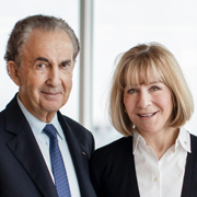 Gerald Schwartz and Heather Reisman smile while standing before a window.